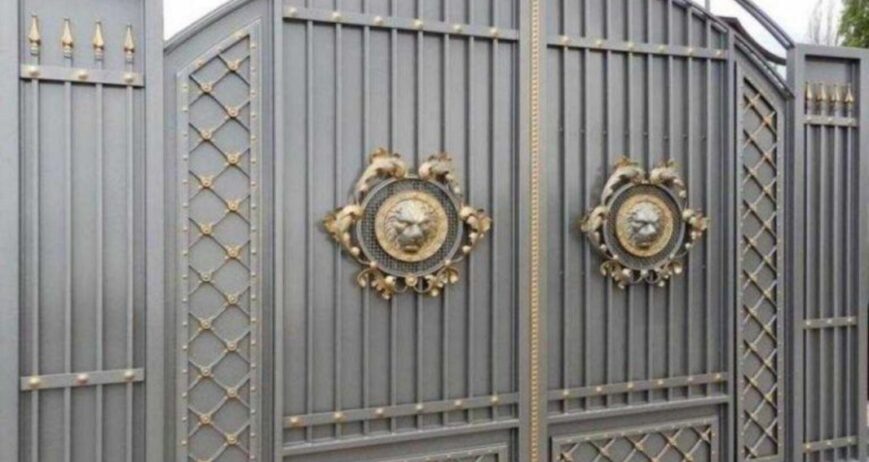A grand, heavy-duty metal driveway gate featuring an arched top frame, intricate lattice side panels, golden finial spikes, and prominent decorative lion head metallic emblems at the center.