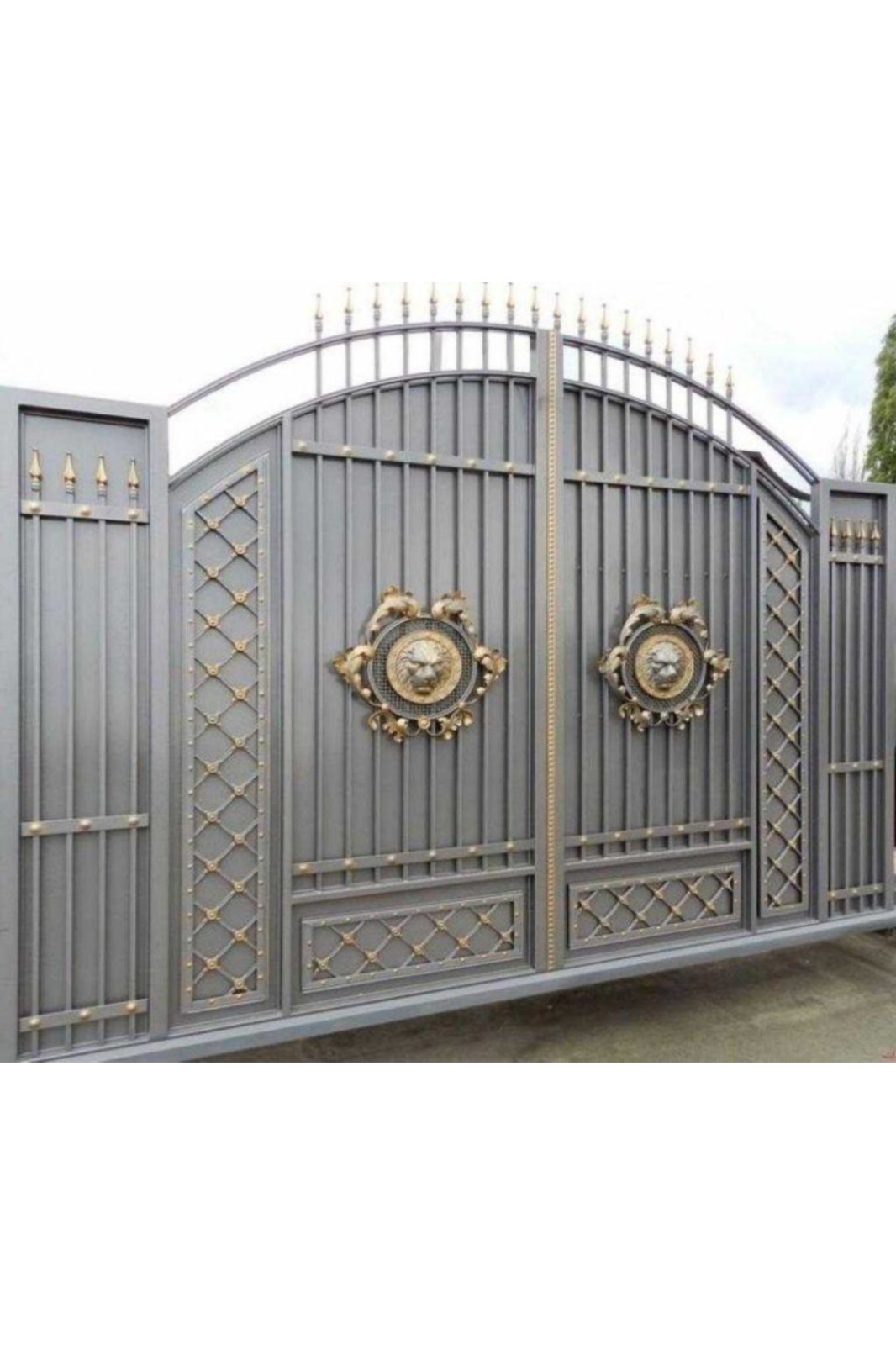 A grand, heavy-duty metal driveway gate featuring an arched top frame, intricate lattice side panels, golden finial spikes, and prominent decorative lion head metallic emblems at the center.